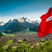 Landschaftsbild von einer Stadt im Tal und Berge und ein See im Hintergrund. Im Vordergrund eine schweizer Flagge. Dieses Bild wurde mit KI generiert.